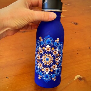 May include: A blue water bottle with a mandala design painted on it. The mandala is made up of blue, white, and gold dots.