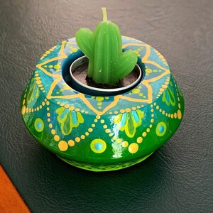 May include: A green and gold painted wooden candle holder with a small green succulent plant in the center. The holder has a mandala design with yellow dots and green and blue floral patterns.