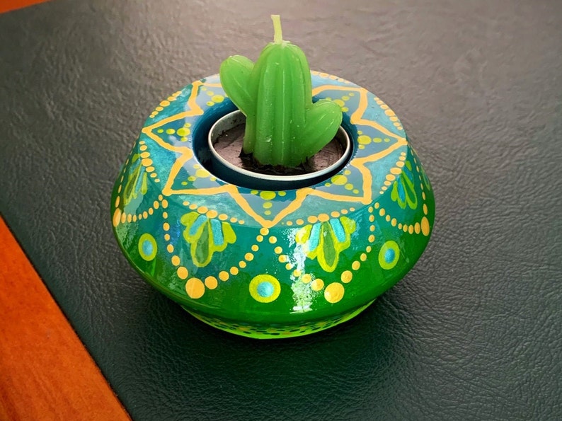 May include: A green and gold painted wooden candle holder with a small green succulent plant in the center. The holder has a mandala design with yellow dots and green and blue floral patterns.