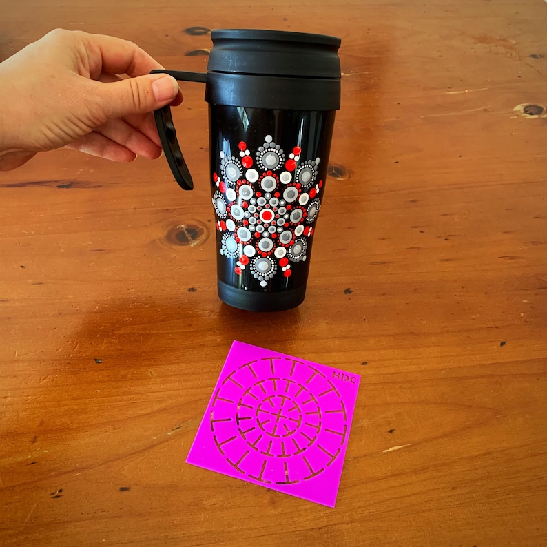 May include: A black travel mug with a red, white, and gray mandala design. A pink plastic stencil with a circular pattern is also shown.