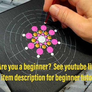 May include: A close-up of a black canvas with a white grid pattern. The center of the canvas features a pink and gold dot mandala design. A hand is holding a red tool and placing a white dot on the canvas. "Are you a beginner? See youtube link in item description for beginner tutorial" is written in yellow text at the bottom of the canvas.