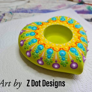 May include: A heart-shaped candle holder with a green base and a mandala design in yellow, orange, blue, and purple. The holder has a small hole in the center for a candle.