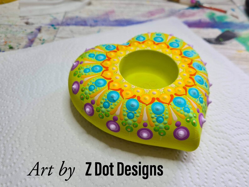 May include: A heart-shaped candle holder with a green base and a mandala design in yellow, orange, blue, and purple. The holder has a small hole in the center for a candle.