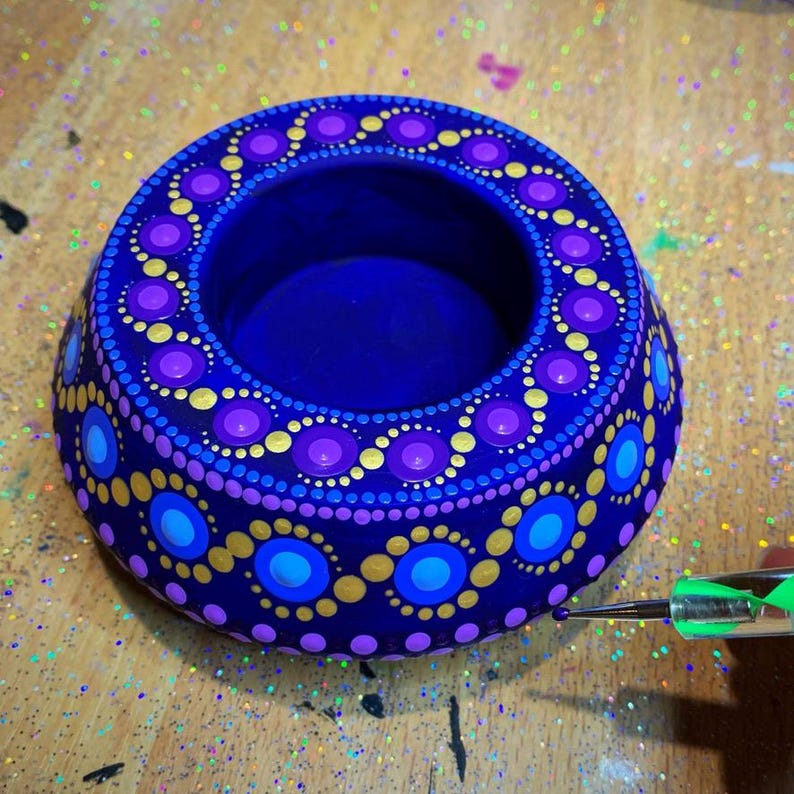 May include: A blue, purple, and gold hand-painted candle holder with a dot mandala design. The holder is being decorated with a dotting tool.