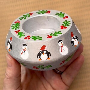 May include: A silver candle holder with a festive winter theme. It features a wreath of green leaves and red berries around the top, and penguins and snowmen painted on the sides.