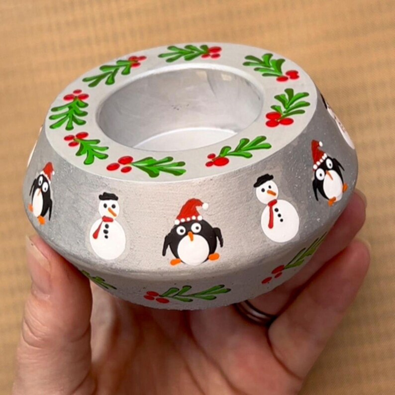 May include: A silver candle holder with a festive winter theme. It features a wreath of green leaves and red berries around the top, and penguins and snowmen painted on the sides.