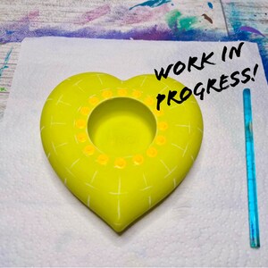 May include: A yellow heart-shaped ceramic planter with a white grid pattern and yellow dots around the rim. The planter is on a white surface with the text "WORK IN PROGRESS!" written in black marker.