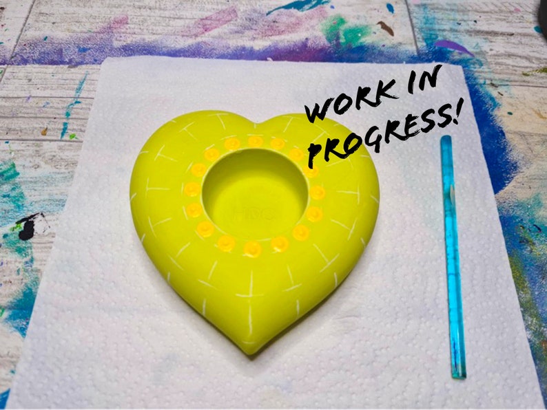 May include: A yellow heart-shaped ceramic planter with a white grid pattern and yellow dots around the rim. The planter is on a white surface with the text "WORK IN PROGRESS!" written in black marker.