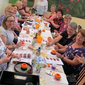 May include: A group of people are gathered around a table, painting ceramic planters. The table is covered in a white tablecloth and is decorated with paintbrushes, paint, and other supplies. The planters are various colors and designs.