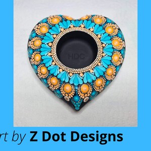 May include: A heart-shaped teal and orange mandala candle holder. The holder is made of black clay and has a circular opening in the center. The mandala design is intricate and detailed, with a variety of colors and patterns.