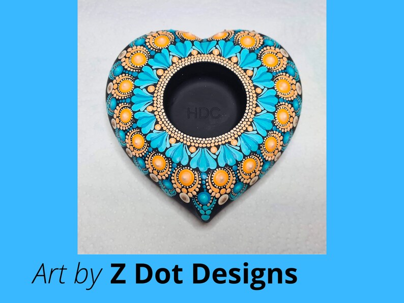 May include: A heart-shaped teal and orange mandala candle holder. The holder is made of black clay and has a circular opening in the center. The mandala design is intricate and detailed, with a variety of colors and patterns.