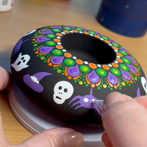 K&ouml;nnte beinhalten: Ein schwarzer, donutf&ouml;rmiger Kerzenhalter aus Keramik mit einem Mandala-Design in Lila, Gr&uuml;n, Orange und Wei&szlig;. Der Halter ist mit Halloween-Motiven verziert, darunter ein Geist, ein Hexenhut, ein Sch&auml;del und eine Spinne.