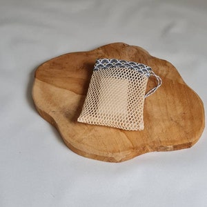 Peut inclure: Un sac en maille beige avec une bordure à motifs blancs et noirs posé sur une surface en bois. Le sac est conçu pour contenir un savon.
