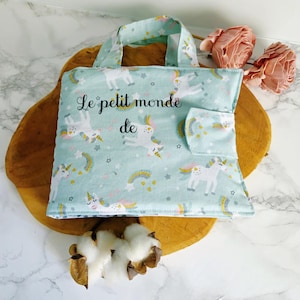 Pode incluir: Uma sacola azul clara com um padrão de unicórnio e o texto "Le petit monde de". A sacola tem um bolso e alças combinando. A sacola está em uma superfície de madeira.