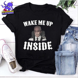 Pode incluir: Camiseta preta com o texto "WAKE ME UP INSIDE" em fonte gótica branca. A camiseta apresenta uma imagem de um homem usando um fone de ouvido. A camiseta é combinada com jeans azuis e tênis brancos.