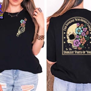 Könnte beinhalten: Schwarzes T-Shirt mit einem Totenkopf-Motiv. Der Totenkopf ist von Blumen umgeben und dem Text "Finde jemanden, der Blumen in den dunkelsten Teilen von dir wachsen lässt".
