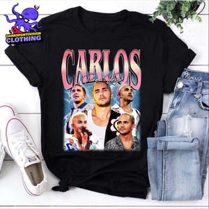 May include: Black t-shirt with a retro design featuring Carlos PenaVega. The design includes the name "CARLOS" in pink and white, and multiple images of the celebrity. The shirt is displayed with blue denim jeans and white trainers.