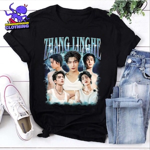 May include: Black t-shirt featuring a graphic of five men with the text "ZHANG LINGHE" in a blue, stylized font. The shirt has a crew neck and short sleeves. The shirt is paired with blue jeans and white sneakers.