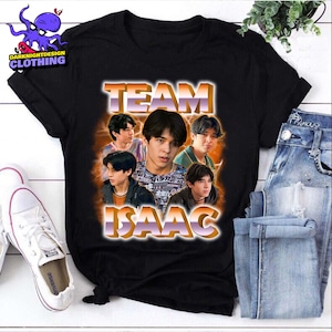 以下が含まれることがあります： 若い男性のグラフィックと「TEAM ISAAC」の文字がオレンジと白のグラデーションで描かれた黒いTシャツ。シャツは青いジーンズと白いスニーカーと組み合わせて表示されています。