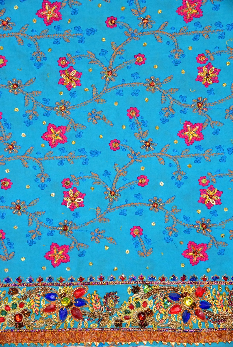 May include: A turquoise fabric with a floral pattern embroidered with pink, yellow, and blue thread. The fabric has a gold and silver trim with colorful jewels.