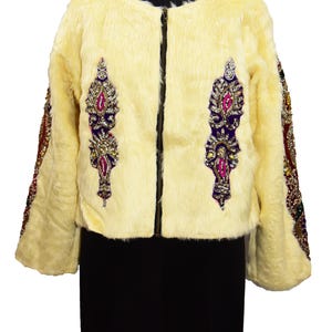 Könnte beinhalten: Cremefarbene Kunstfelljacke mit schwarzem Reißverschluss. Die Jacke hat verzierte, juwelenfarbene Stickereien auf der Vorderseite und den Ärmeln. Die Ärmel sind mit bunten Perlen verziert. Die Jacke wird auf einer schwarzen Schaufensterpuppe präsentiert.