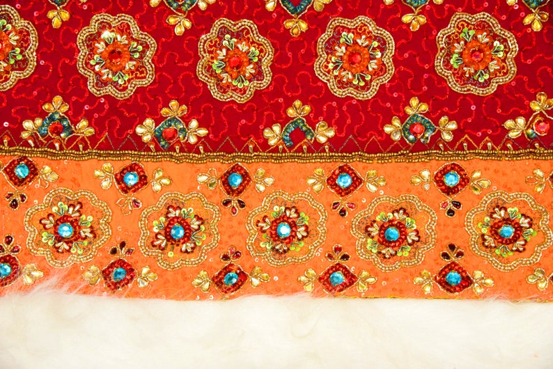 May include: A close-up of a red and orange fabric with intricate embroidery. The fabric has a beaded border and a white fur trim.