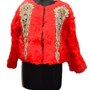 Könnte beinhalten: Eine leuchtend rote Jacke mit flauschiger Textur. Die Jacke ist mit aufwendigen Verzierungen mit goldenen, silbernen, blauen und roten Akzenten auf der Vorderseite versehen. Sie hat einen durchgehenden Reißverschluss und wird auf einer schwarzen Schaufensterpuppe präsentiert.