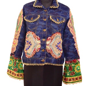 Könnte beinhalten: Eine blaue Jeansjacke mit Tarnmuster und verziert mit goldenen Perlen, Pailletten und bunten Stickereien. Die Jacke hat einen Knopfverschluss und zwei Taschen.
