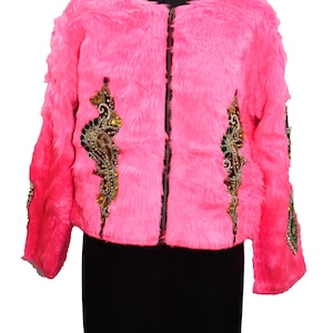 Könnte beinhalten: Eine leuchtend pinkfarbene Kunstfelljacke mit Reißverschluss. Die Jacke ist mit aufwendigen Verzierungen mit farbigen Edelsteinen und goldfarbenen Akzenten auf der Vorderseite und den Ärmeln versehen. Sie wird auf einer schwarzen Schneiderpuppe präsentiert.