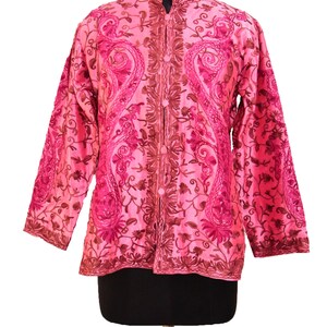 Puede incluir: Una chaqueta rosa bordada con cuello mandarín y cierre de botones. La chaqueta presenta un diseño floral y paisley en tonos rosa y marrón.