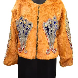 Könnte beinhalten: Eine Kunstfelljacke in einem warmen Orangeton mit aufwendigen Verzierungen. Die Jacke hat einen Reißverschluss vorne und ist mit kunstvollen, juwelenfarbenen Designs in Blau, Rot und Gold verziert. Die Ärmel haben ebenfalls dekorative Details. Die Jacke wird auf einer schwarzen Schaufensterpuppe präsentiert.