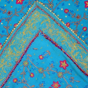 May include: A turquoise fabric with a gold, pink, and blue embroidered floral pattern. The fabric has a gold trim around the edges.