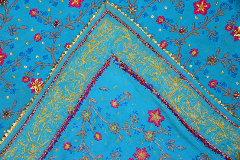 May include: A turquoise fabric with a gold, pink, and blue embroidered floral pattern. The fabric has a gold trim around the edges.