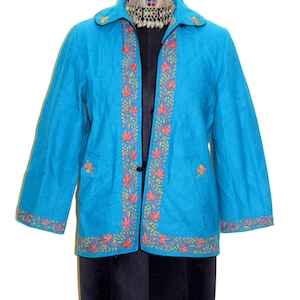 Puede incluir: Chaqueta turquesa con bordados florales naranjas y amarillos en los bordes y bolsillos. La chaqueta tiene un botón negro y se exhibe en un maniquí negro. La chaqueta es de longitud media. Medidas en centímetros.