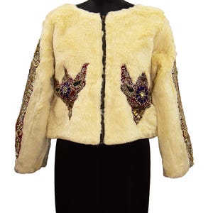 Könnte beinhalten: Cremefarbene Kunstfelljacke mit schwarzem Reißverschluss. Die Jacke hat verzierte, farbenfrohe Perlenverzierungen auf Brust und Ärmeln. Die Ärmel sind mit goldenen und roten Perlen besetzt. Die Jacke wird auf einer schwarzen Schneiderpuppe präsentiert.