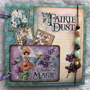 Può includere: Un album di ritagli quadrato con una copertina a motivi color verde acqua, decorato con illustrazioni di fate, fiori e il testo "FAIRIE DUST" e "Believe in MAGIC". Il libro è rilegato con anelli metallici viola e un cordino verticale con perline.
