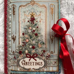 Puede incluir: Un diario vintage con temática navideña con una cubierta azul claro que presenta un árbol de Navidad decorado y la palabra "GREETINGS". El diario tiene una cinta roja y un lomo rojo decorativo.