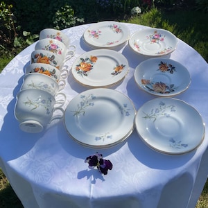 May include: A vintage tea set with floral patterns. The cups and saucers feature a variety of floral designs, including roses and bluebells. The set is arranged on a white tablecloth.