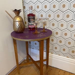 May include: A gold pineapple cocktail shaker, a bottle of flavored liquor, and two stemmed glasses are on a small, round purple table with wooden legs. The table is in front of patterned wallpaper.