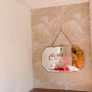 May include: An octagonal wall mirror with a gold-toned frame, suspended by a metal chain. The glass features an etched decorative floral design and the text "LES ROSES DE SAADI" in the centre. The mirror reflects a red object and a wooden door.