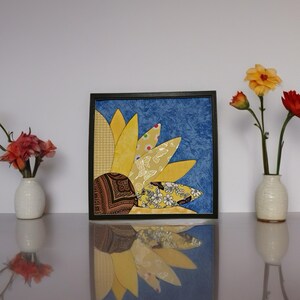May include: A framed fabric art piece featuring a sunflower design. The sunflower has yellow petals with patterned fabric accents against a blue background. The artwork is displayed on a reflective surface, with vases of flowers on either side.