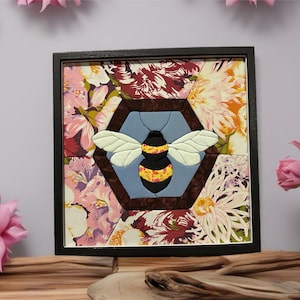 May include: Framed bee-themed artwork. The bee is centered within a hexagonal shape, with a black and yellow body and white wings. The background features a floral pattern in shades of pink, white, and burgundy. The artwork is encased in a black frame.