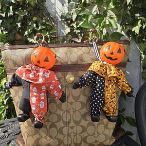 May include: Two pumpkin-headed dolls hanging from a brown monogrammed tote bag. The left doll wears a black and orange coffee-themed outfit, while the right doll wears a black polka-dot and candy corn patterned outfit with ruffled collars.
