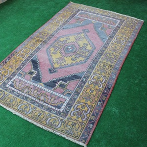 K&ouml;nnte beinhalten: Ein antiker t&uuml;rkischer Teppich mit einem geometrischen Muster in Rosa, Gelb und Blau. Der Teppich hat einen Fransenrand und liegt auf einer gr&uuml;nen Oberfl&auml;che.