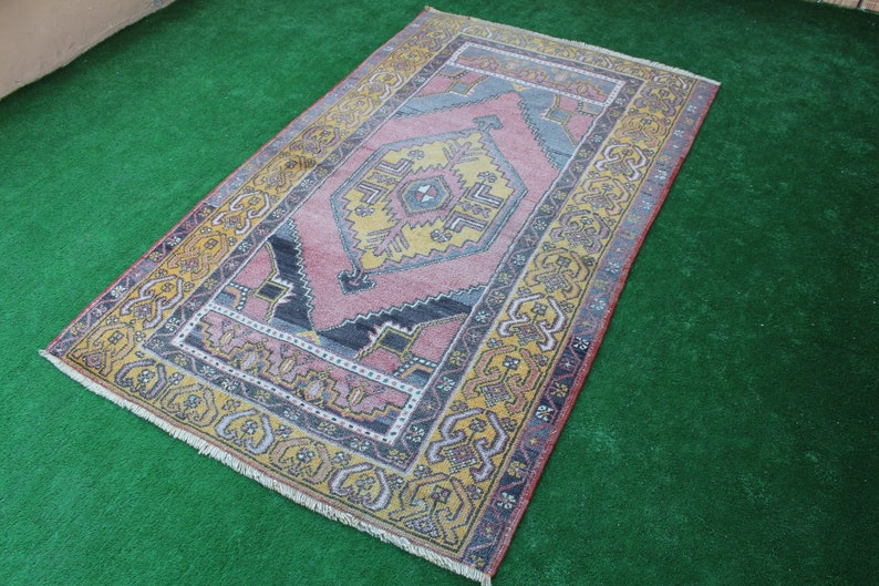 K&ouml;nnte beinhalten: Ein antiker t&uuml;rkischer Teppich mit einem geometrischen Muster in Rosa, Gelb und Blau. Der Teppich hat einen Fransenrand und liegt auf einer gr&uuml;nen Oberfl&auml;che.