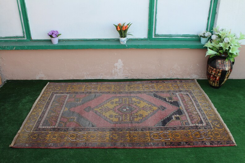 K&ouml;nnte beinhalten: Ein antiker t&uuml;rkischer Teppich mit einem geometrischen Muster in Rosa, Gelb und Blau. Der Teppich hat einen Fransenrand und liegt auf einer gr&uuml;nen Oberfl&auml;che.