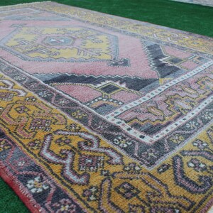 K&ouml;nnte beinhalten: Ein Vintage-Teppich mit einem geometrischen Muster in Rosa, Gelb und Grau. Der Teppich hat einen Fransenrand und ist aus Wolle.