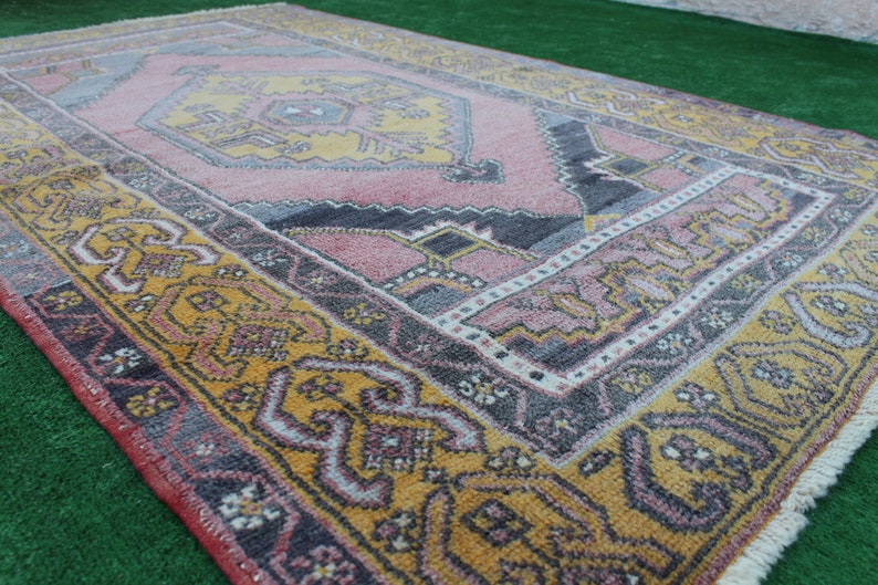 K&ouml;nnte beinhalten: Ein Vintage-Teppich mit einem geometrischen Muster in Rosa, Gelb und Grau. Der Teppich hat einen Fransenrand und ist aus Wolle.