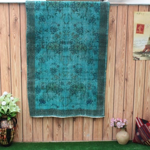 Pu&ograve; includere: Un tappeto vintage blu turchese con un motivo floreale &egrave; appeso a una parete di legno. Il tappeto ha un bordo verde scuro.