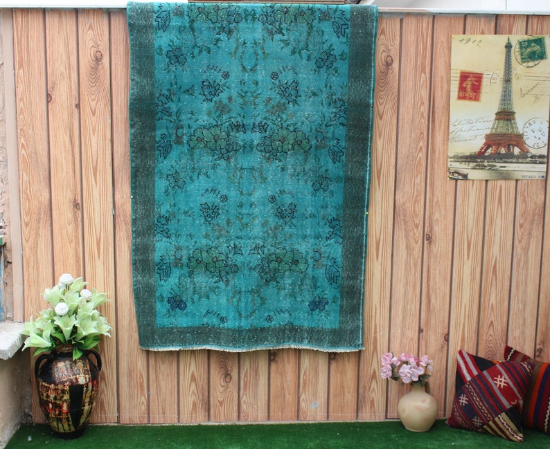 Pu&ograve; includere: Un tappeto vintage blu turchese con un motivo floreale &egrave; appeso a una parete di legno. Il tappeto ha un bordo verde scuro.